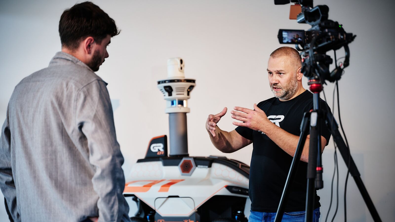 A man in a GENERATIVE Robotics T-shirt gives a camera interview next to the GR-01, a television camera on a tripod in front of him